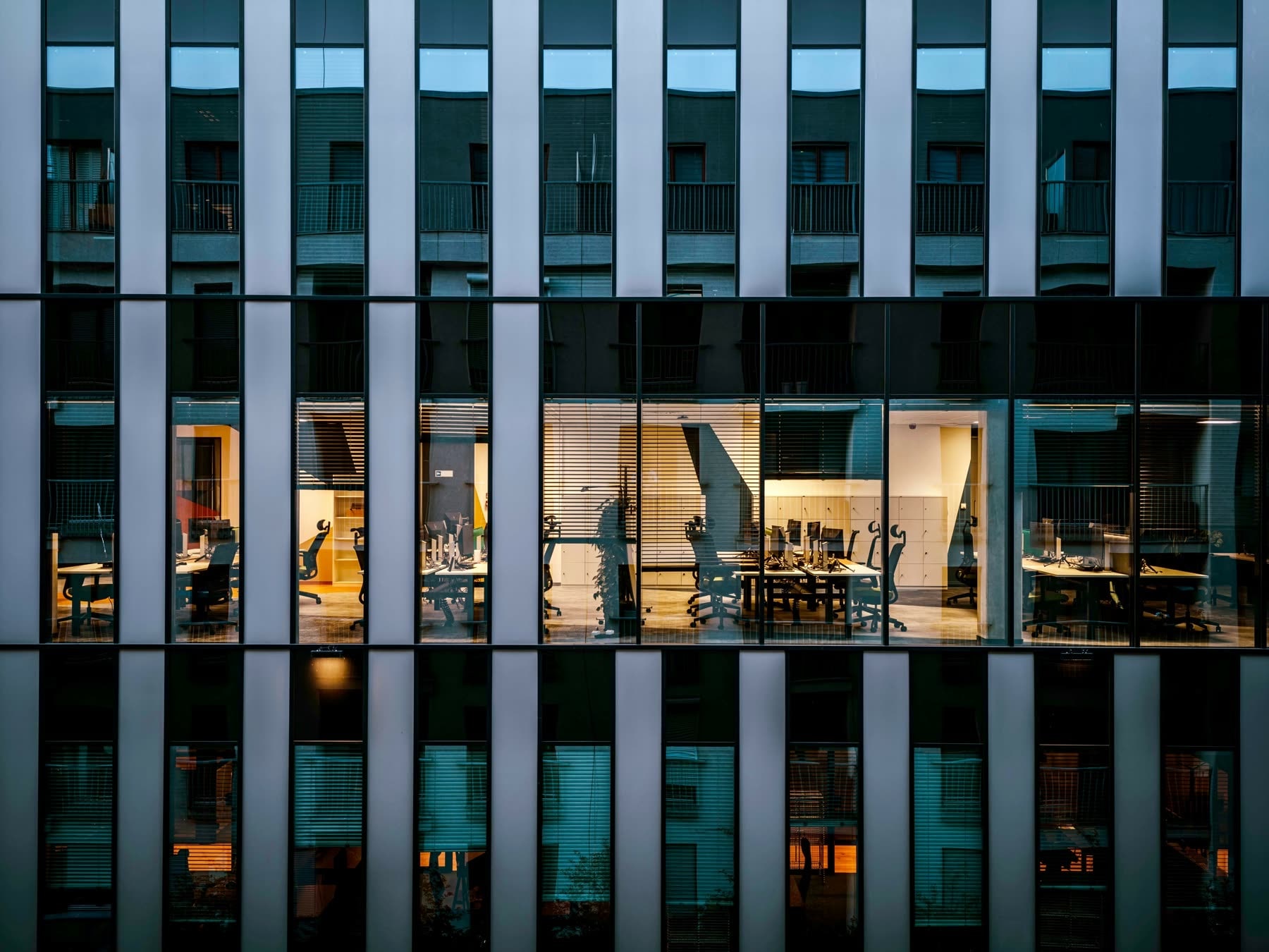 Lit office floors seen through the glass and steel grid of a building at dusk.