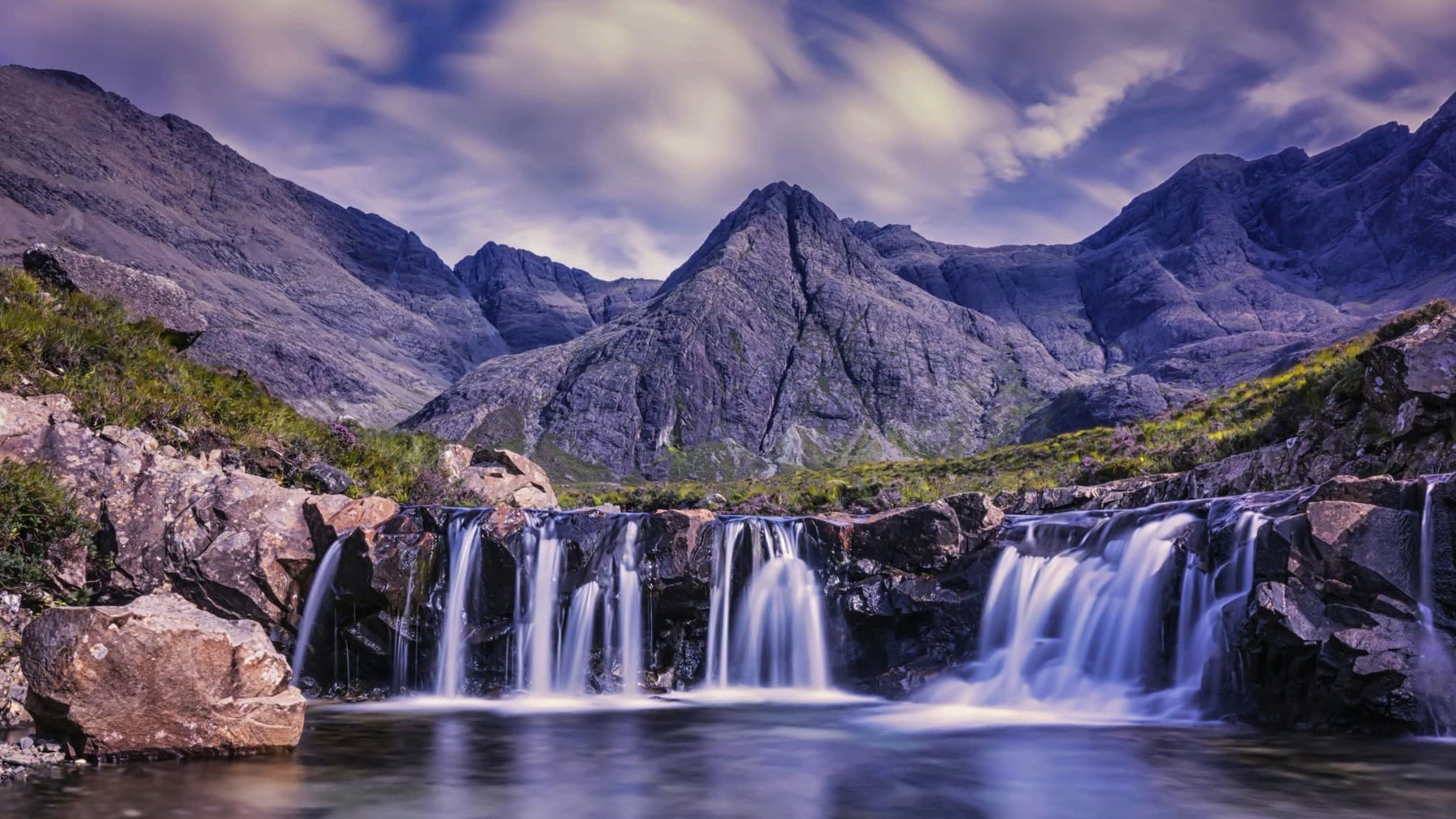 A wide waterfall pouring over dark rocks into a clear pool, with jagged mountains under a swirling sky.